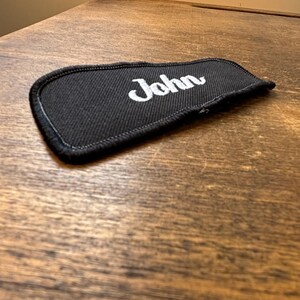 以下が含まれることがあります： 白い筆記体で「John」と刺繍された黒い布パッチ。 パッチには黒い縁取りがあり、木製の表面に表示されています。