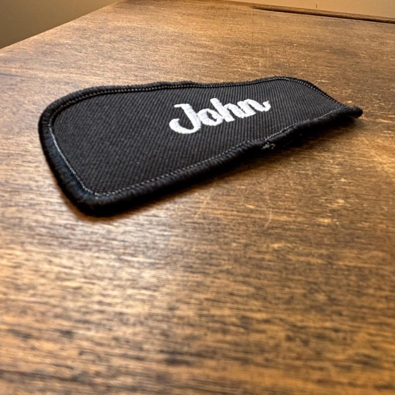 以下が含まれることがあります： 白い筆記体で「John」と刺繍された黒い布パッチ。 パッチには黒い縁取りがあり、木製の表面に表示されています。