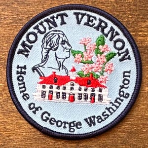 May include: A circular embroidered patch with a light blue background and a dark blue border. It features a profile of George Washington, a white building with a red roof, and pink flowers. The text reads Mount Vernon Home of George Washington.