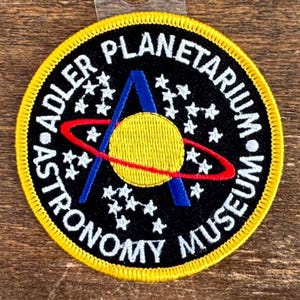 Puede incluir: Parche bordado con el texto "Adler Planetarium Astronomy Museum" en blanco sobre fondo negro. El parche presenta un círculo amarillo, un anillo rojo y un triángulo azul. Un borde amarillo rodea el parche.