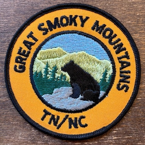 May include: Embroidered patch with a circular design featuring a black bear, mountains, and pine trees. The text "GREAT SMOKY MOUNTAINS" and "TN/NC" are in black lettering on a yellow background with a black border.