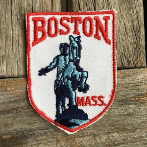 May include: Embroidered patch featuring the word "BOSTON" in red, arched over a blue and white statue of a person on a horse. The word "MASS" is below the statue, all on a white background with a red border.