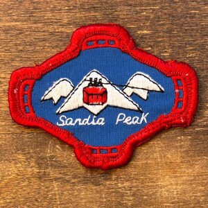 May include: Embroidered patch featuring a blue background with a white mountain range and a red cable car. The patch has a red border and the words "Sandia Peak" in white script.