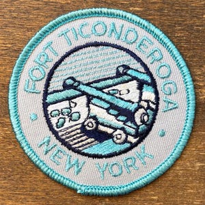 May include: Round patch with the text "FORT TICONDEROGA NEW YORK" in teal embroidery. The patch features a teal and white illustration of a cannon and a building. The patch has a light gray background and a teal border.