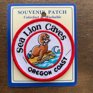 May include: A red, white, and blue embroidered patch with the text "Sea Lion Caves Oregon Coast". A cartoon sea lion is depicted on a rock in the ocean.