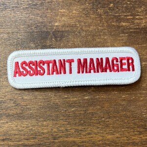 May include: A rectangular embroidered patch with a white background and a white stitched border. The text ASSISTANT MANAGER is embroidered in bold red capital letters across the center of the patch, featuring a clean and legible font style.