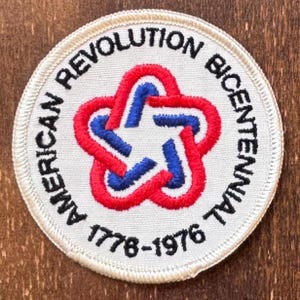 Peut inclure: Écusson circulaire avec le texte "AMERICAN REVOLUTION BICENTENNIAL" en noir sur le bord. Un motif en forme d'étoile rouge et bleu est au centre. Les années "1776-1976" sont imprimées en bas. L'écusson a un fond blanc et une bordure crème.