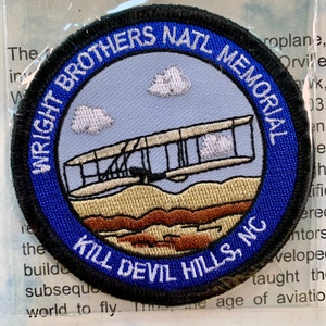 May include: A blue and black embroidered patch with the text "Wright Brothers Natl Memorial Kill Devil Hills, NC". The patch features a white aeroplane flying over a brown landscape with white clouds in the sky.