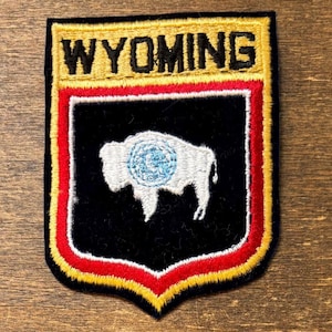 Puede incluir: Parche bordado con la palabra "WYOMING" en oro sobre un escudo negro. El escudo presenta un bisonte blanco con un centro azul y blanco. El parche está bordeado con costuras rojas y doradas.