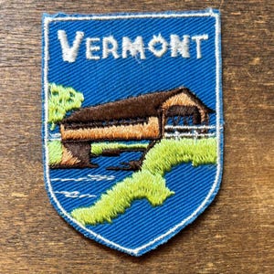 以下が含まれることがあります： 青い背景に白い大文字でVERMONTと書かれた盾形の刺繍ワッペン。茶色の屋根付き橋が青い小川にかかっており、鮮やかな緑色の草地と小さな木が描かれています。