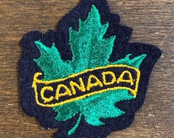 Puede incluir: Un parche bordado que presenta una hoja de arce de color verde vibrante centrada sobre un fondo azul marino oscuro. Una banda horizontal amarilla está cosida en el centro de la hoja, con la palabra CANADA en letras azul marino oscuras.