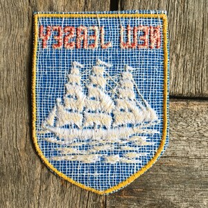 May include: A shield-shaped embroidered patch with a blue background and a yellow border. The patch features a white sailing ship graphic and the red embroidered text NEW JERSEY, which appears mirrored or reversed in the design.