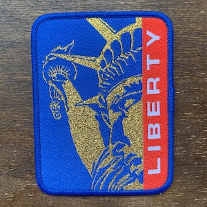 May include: A blue and red embroidered patch with a gold outline of the Statue of Liberty and the word "LIBERTY" in white letters.