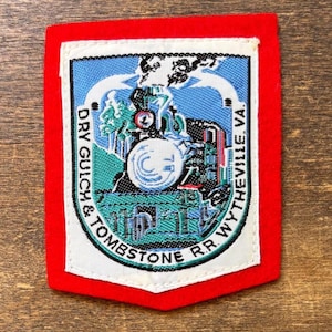 May include: A rectangular embroidered patch with a bright red border and a white inner frame. The center features a blue and green graphic of a steam locomotive with smoke, surrounded by the text DRY GULCH & TOMBSTONE R.R. WYTHEVILLE, VA.
