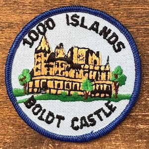 Op de afbeelding: Geborduurde patch met Boldt Castle van de 1000 Islands. De patch heeft een lichtblauwe achtergrond met een donkerblauwe rand. Het kasteel is afgebeeld in goud en zwart, met groene bomen en de tekst "1000 ISLANDS" en "BOLDT CASTLE".
