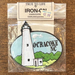Puede incluir: Parche bordado con un faro blanco con el texto "OCRACOKE NC" sobre un fondo azul claro y verde. El parche está etiquetado como "IRON-ON" y "EMBROIDERE EMBLEMS/patches".