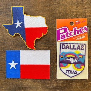 May include: Three iron-on patches featuring Texas themes. One patch is the shape of Texas with a blue, white, and red design. Another is a rectangular Texas flag. The third patch is a Dallas, Texas design.