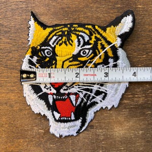 May include: An embroidered tiger head patch featuring yellow, black, and white fur details with a red open mouth and sharp white teeth. A white measuring tape is placed across the patch, showing a width of approximately 3.5 inches.
