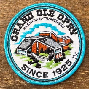 May include: Embroidered patch featuring the Grand Ole Opry in Nashville, Tennessee. The patch has a circular design with a teal border and the words "Grand Ole Opry" and "Since 1925". The image depicts the Opry building with orange and gray colors.