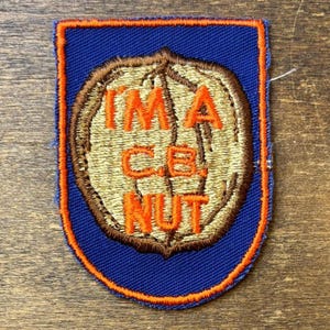Könnte beinhalten: Ein schildförmiger, bestickter Aufnäher mit blauem Hintergrund und orangefarbenem Rand. In der Mitte befindet sich eine beige Walnussgrafik mit dem orangefarbenen Sticktext "I'M A C.B. NUT", der in drei Zeilen auf der Nuss angeordnet ist.