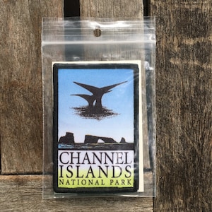 Op de afbeelding: Een zwart-witte patch met een blauwe achtergrond. De patch toont een silhouet van twee vogels die over een rotsachtig eiland vliegen. De tekst "CHANNEL ISLANDS NATIONAL PARK" is in wit op een zwarte achtergrond gedrukt.