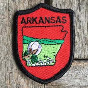Pode incluir: Patch bordado em forma de escudo com o estado do Arkansas. O patch tem um fundo vermelho com uma borda preta. O contorno do estado é verde e apresenta uma planta de algodão e a palavra "ARKANSAS".