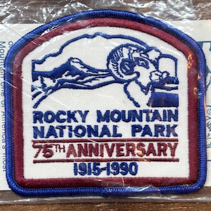 Op de afbeelding: Een rechthoekige patch met een blauwe rand en een witte achtergrond. De patch toont een blauwe illustratie van een berggeit en een berglandschap. Tekst op de patch luidt "ROCKY MOUNTAIN NATIONAL PARK" en "75TH ANNIVERSARY 1915-1990".