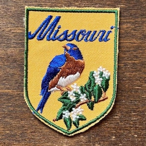 May include: Embroidered patch featuring a bluebird perched on a flowering branch, set against a yellow background. The word "Missouri" is arched above the bird in blue script. The patch has a green border.