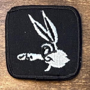 May include: A square-shaped black embroidered patch featuring a white stitched graphic of a cartoon rabbit head in profile, with long ears pointing upward and a fluffy tail, finished with a black stitched border around the entire perimeter.