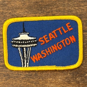 May include: Embroidered patch featuring the Seattle Space Needle in black, white, and silver against a blue background. The text "SEATTLE WASHINGTON" is in orange. The patch has a yellow border.