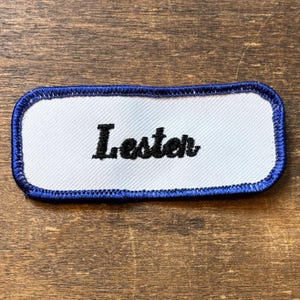 Peut inclure: Un écusson rectangulaire avec le nom "Lester" brodé en noir sur un fond blanc. Le patch a une bordure bleue, probablement pour un uniforme ou un vêtement de travail.