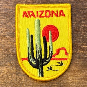 Pode incluir: Um emblema bordado em forma de escudo com fundo amarelo. O texto ARIZONA está bordado em vermelho no topo. Um cacto saguaro verde encontra-se no centro, com um sol vermelho e linhas de paisagem desértica vermelhas ao fundo.