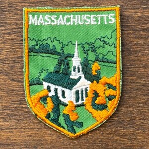 May include: Embroidered patch featuring a church with a steeple, set against a green background. The word "MASSACHUSETTS" is stitched across the top in white. The patch has a gold border and is likely a souvenir or collectible item.