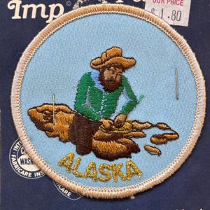 Könnte beinhalten: Ein runder, blauer gestickter Aufnäher mit einem braunen Rand. Der Aufnäher zeigt ein Cartoon-Bild eines Mannes, der ein grünes Hemd, eine braune Hose und einen braunen Hut trägt und nach Gold sucht. Der Text "ALASKA" ist in goldenen Buchstaben unter dem Bild gestickt.