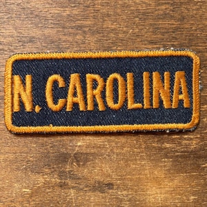 May include: A rectangular patch with the text "N. CAROLINA" embroidered in gold thread on a navy blue background. The patch has a gold border and is set against a wooden surface.