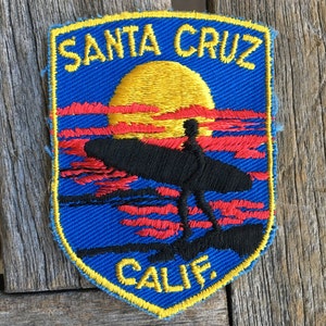 May include: A blue and yellow embroidered patch with the text "SANTA CRUZ CALIF" and a silhouette of a surfer riding a wave at sunset.