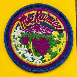 May include: A blue and yellow embroidered patch with the text "Mt. Rainier" in a white outline. The patch features a stylized mountain with white peaks, green grass, purple flowers, and white flowers with yellow centers.