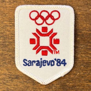 May include: A white patch with a red and blue design. The top features the Olympic rings, with a stylized red emblem below. The text "Sarajevo '84" is in blue. The patch has a white border.