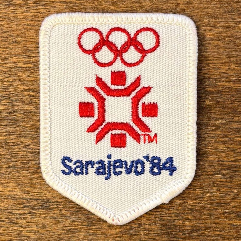 Puede incluir: Un parche blanco con un dise&ntilde;o rojo y azul. La parte superior presenta los anillos ol&iacute;mpicos, con un emblema rojo estilizado debajo. El texto "Sarajevo '84" est&aacute; en azul. El parche tiene un borde blanco.