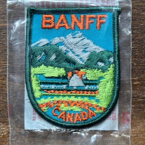 Puede incluir: Parche bordado con la palabra "BANFF" sobre una escena de montaña con un lago y árboles. El parche tiene un fondo turquesa y un borde verde. El texto "ALBERTA CANADA" está en la parte inferior en naranja.