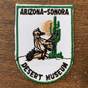 May include: Embroidered patch featuring a roadrunner, cactus, and sun design. The patch has a white background with green trim and the text "ARIZONA-SONORA DESERT MUSEUM". The roadrunner is depicted in shades of brown and black.
