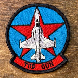May include: Embroidered patch featuring a white jet fighter superimposed over a red star, set against a blue background. The design is encircled by a black border and includes the text "TOP GUN" in red.