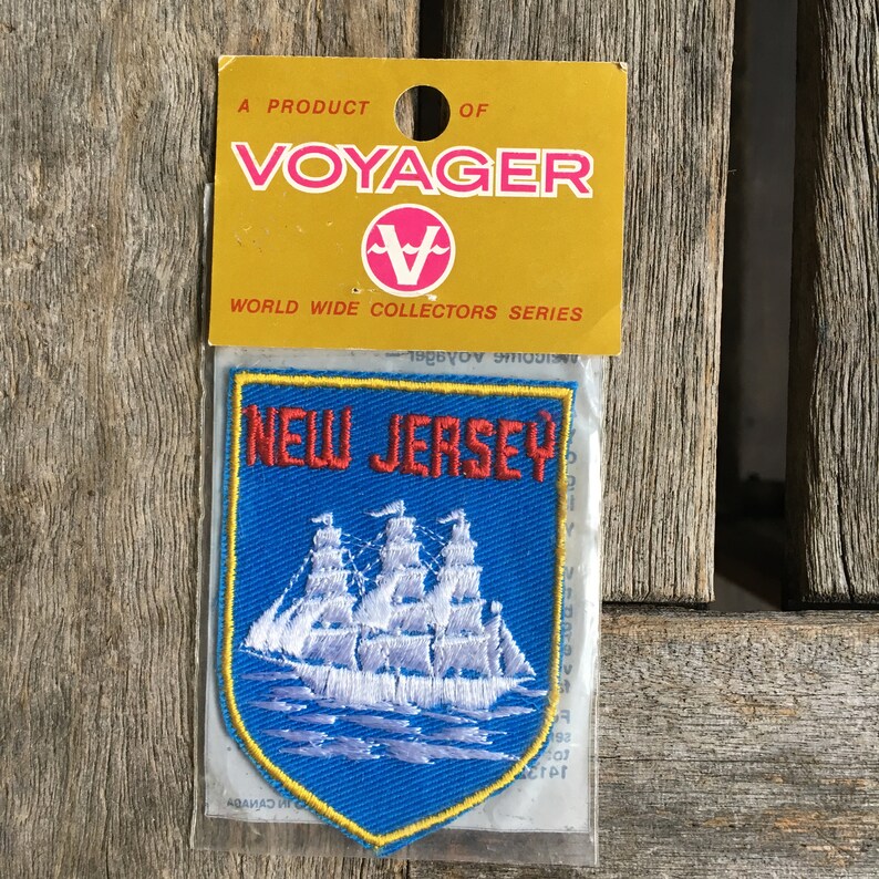 May include: A blue embroidered patch featuring a white sailing ship and the red text NEW JERSEY, enclosed in a yellow border. The patch is attached to a yellow cardboard header card with the text A PRODUCT OF VOYAGER WORLD WIDE COLLECTORS SERIES.