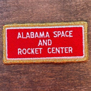 May include: Rectangular embroidered patch featuring the text "ALABAMA SPACE AND ROCKET CENTER" in white on a red field. It has a gold border and a pink inner border. The patch is designed for application to fabric.