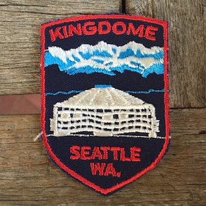 May include: A blue and red embroidered patch with the words "Kingdome Seattle WA" and a white building with a blue roof.