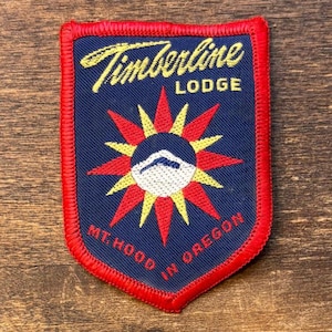 Pode incluir: Um patch Timberline Lodge azul marinho com uma borda vermelha. O patch apresenta um design de explosão solar com uma silhueta de montanha e o texto "Timberline Lodge" e "Mt. Hood in Oregon" em amarelo e vermelho.