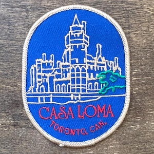 Puede incluir: Parche ovalado con fondo azul y contorno blanco. El parche presenta un dibujo lineal blanco de un castillo con las palabras "CASA LOMA TORONTO, CAN." en rojo. Un diseño verde está en el lado derecho del castillo.