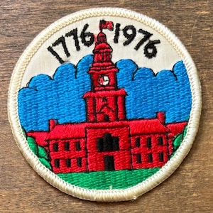 May include: A circular patch featuring a red building with a clock tower, set against a blue sky. The patch is bordered by a white stitched edge, with the years "1776" and "1976" arched above the building in black.
