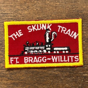 Könnte beinhalten: Ein rechteckiger Aufnäher mit rotem Hintergrund und gelbem Rand. Der Aufnäher zeigt eine schwarz-weiße Zugillustration mit dem Text "THE SKUNK TRAIN" darüber und "FT. BRAGG-WILLITS" darunter.