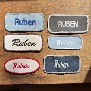 May include: Six rectangular and oval embroidered patches featuring the name Ruben in various fonts and colours. The patches include light blue, dark grey, white, and dark blue fabric bases with stitched borders in matching or contrasting thread colours.